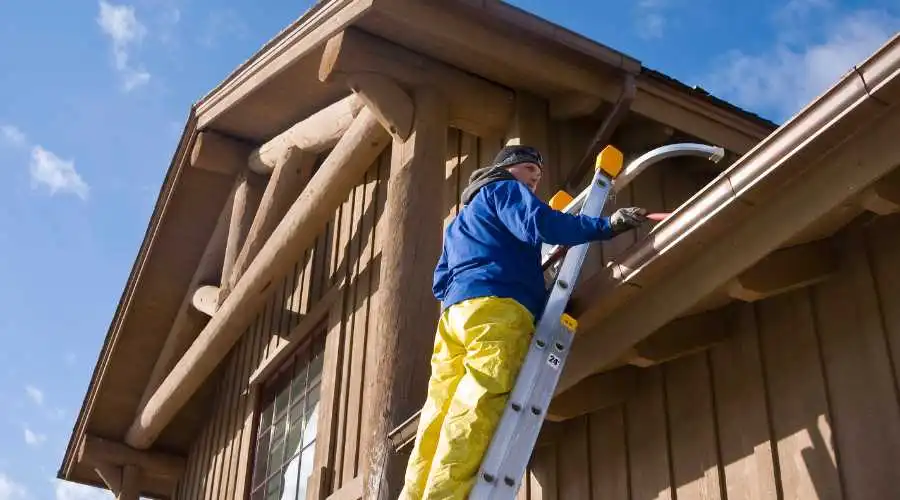 A man in yellow waterproof pants on a ladder cleaning a gutter